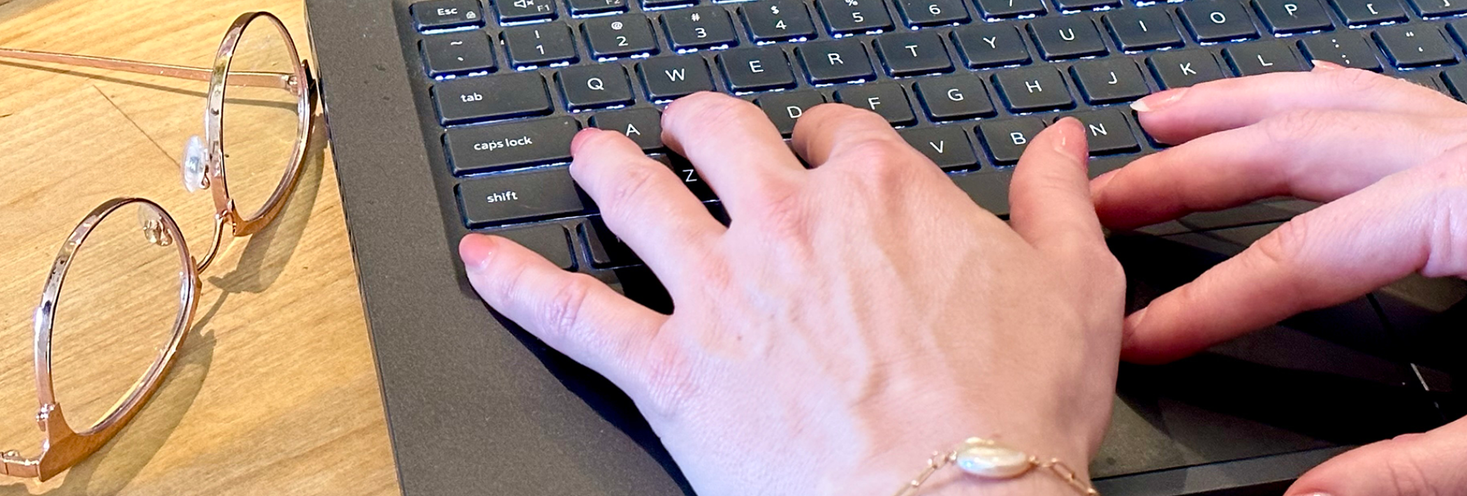 Hands typing on a laptop lying on a desk next to a pair of gold eyeglasses.